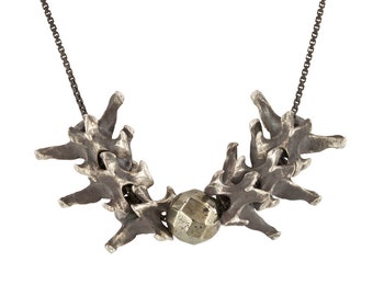 Op de afbeelding: Een zilveren ketting met een hanger gemaakt van verschillende met elkaar verbonden botvormige stukken. De hanger is gecentreerd rond een grote, ronde, gefacetteerde pyriet steen.