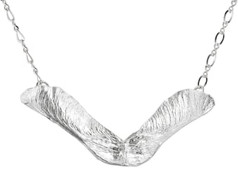 Op de afbeelding: Een zilveren ketting met een hanger in de vorm van een esdoornzaad. De hanger is getextureerd en heeft een delicate ketting.