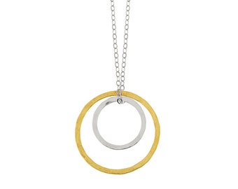 Op de afbeelding: Een gouden en zilveren ketting met een dubbele cirkel hanger. De grotere cirkel is van goud en heeft een gehamerde textuur. De kleinere cirkel is van zilver en zit in de grotere cirkel.