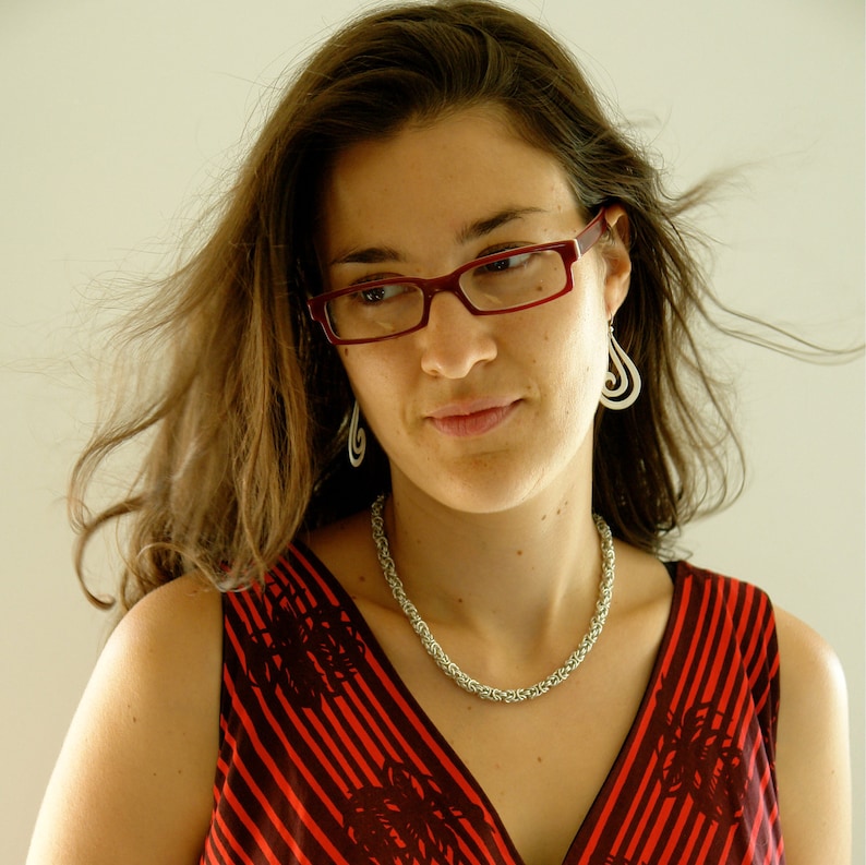 May include: A woman with long brown hair wearing red and black striped clothing. She is wearing red glasses and silver earrings. She is also wearing a silver chain necklace.