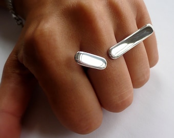 Op de afbeelding: Twee zilveren ringen, één op elke vinger, worden getoond op de hand van een persoon. De ringen zijn beide rechthoekig van vorm en hebben een gladde, gepolijste afwerking.