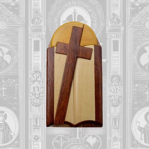 May include: A wooden puzzle box with a cross design. The box is made of light and dark wood and has a hinged lid. The cross is made of a darker wood and is set against a lighter wood background.