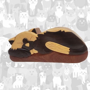 May include: A wooden cat-shaped box in shades of brown, tan, and black. The box is designed to resemble a sleeping cat, with its paws tucked in and head resting on its body. The background features a pattern of various cat silhouettes.