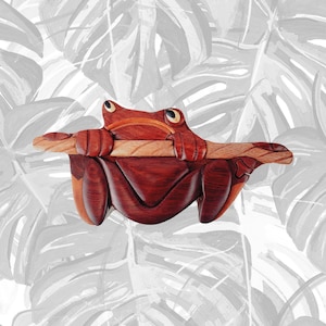May include: A handcrafted wooden frog sculpture in shades of reddish-brown and light brown. The frog is holding a wooden log, with its eyes open. The sculpture is set against a backdrop of gray and white leaf patterns.