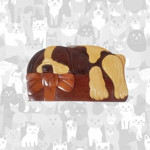May include: A wooden box shaped like a sleeping dog, with a brown body and tan accents. The dog rests on a rectangular base with a bow. The background features a pattern of cat silhouettes.