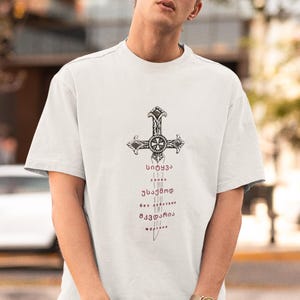 Op de afbeelding: Lichtgrijs t-shirt met een gedetailleerd kruisontwerp en gestileerde tekst. Het shirt met korte mouwen heeft een relaxte pasvorm. De persoon draagt een zwarte pet en zonnebril. Het horloge heeft een goudkleurige wijzerplaat.