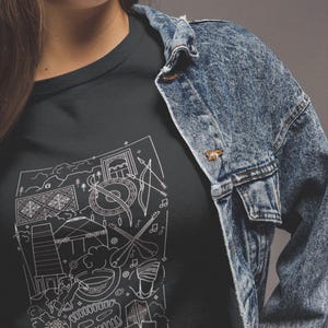 May include: Dark gray t-shirt featuring a white line drawing of cultural elements, including musical instruments, buildings, and decorative patterns. The shirt is worn with a blue denim jacket.