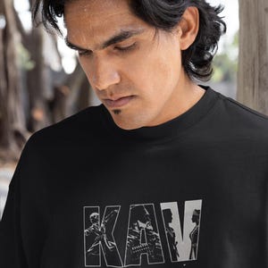 May include: Black t-shirt with the word "KAV" in a white, outlined font. The letters are filled with grayscale images of people. The shirt is worn by a person with dark hair.