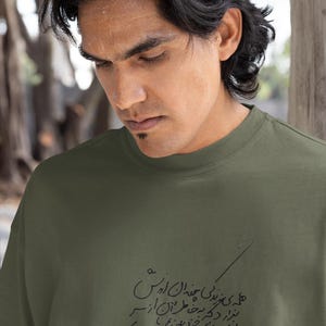 May include: Olive green crew neck t-shirt with black script. The text appears to be in a foreign language. The shirt is made of a soft, comfortable material.