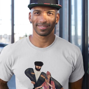 May include: A light gray t-shirt featuring a colorful illustration of two people in traditional attire. The person in the image is wearing a black cap with a green brim and black earrings.