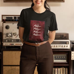 Könnte beinhalten: Schwarzes T-Shirt mit einem rechteckigen Design mit weißer Kalligraphie auf burgunderrotem Hintergrund. Die Person im Bild trägt eine braune Hose und einen braunen Gürtel. Der Hintergrund umfasst Vintage-Audiogeräte und ein dekoratives Regal.