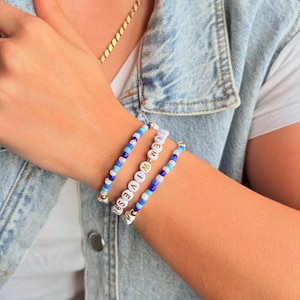 May include: Three beaded bracelets in shades of blue and white. One bracelet spells out "NEW WIVES" in gold letters. The bracelets are worn on a wrist, with a gold chain visible in the background. The bracelets are a fun accessory.