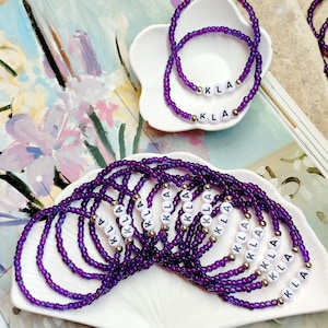 May include: A collection of purple beaded bracelets featuring white letter beads that spell out KLA, accented with small metallic gold beads. The bracelets are arranged on two white, flower-shaped ceramic dishes, showcasing the uniform design.