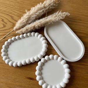 Op de afbeelding: Drie witte keramische dienbladen. Twee van de dienbladen zijn rond met een geperlde rand. Het derde dienblad is ovaalvormig met een gladde rand.
