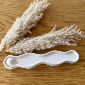 Könnte beinhalten: Ein weißer, wellenförmiger Räucherstäbchenhalter mit einer kleinen Kugel an einem Ende. Zwei beige Pampasgrasstiele sind hinter dem Halter auf einer Holzoberfläche positioniert. Der Halter ist zum Auffangen von Asche konzipiert.