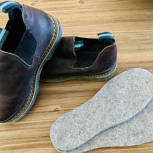 May include: A pair of brown leather slip-on shoes with elastic side panels and a blue and teal stripe detail on the heel. Gray felt insoles are next to the shoes on a wooden surface. The shoes have a stitched sole.