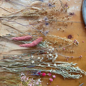 May include: A collection of dried flowers and grasses, including lavender, wheat, and pink wildflowers, arranged on a wooden surface. A pair of gold scissors is also visible.