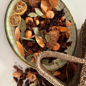 May include: An oval, vintage-style tray with a green and white checkered pattern, filled with dried botanicals. The arrangement includes dried orange slices, bay leaves, and various brown and tan dried flowers and seed pods. A woven basket handle is visible.