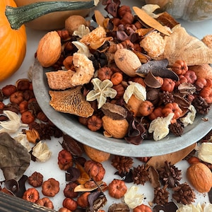 May include: A rustic arrangement featuring a metal dish filled with dried potpourri, including small orange pumpkins, nuts, and various dried flowers and seed pods. The arrangement is set against a backdrop of pumpkins in shades of orange and tan.