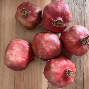 May include: Six whole pomegranates with deep red, leathery skins and textured surfaces. Each fruit features a prominent, dried, star-shaped calyx at the top. The pomegranates are arranged in a cluster, displaying varied shades of crimson and maroon.