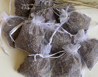 May include: A group of ten small white mesh bags filled with dried lavender flowers. The bags are tied with white ribbon and are arranged on a white surface.