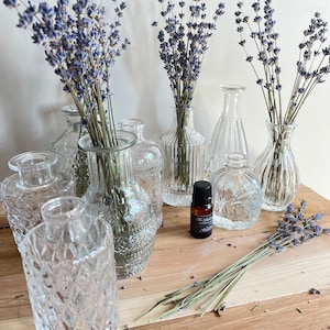 May include: A collection of clear glass bud vases with dried lavender stems. The vases have various textures and shapes, including ribbed, patterned, and smooth designs. A small essential oil bottle is also present on the wooden surface.