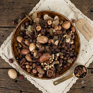 May include: A round brown ceramic bowl filled with a mixture of dried botanicals, including nuts, seed pods, and leaves. A small metal scoop contains a portion of the potpourri, and a tan tag reads Handmade Potpourri - 1 LB.