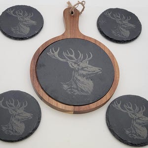 May include: A set of five slate coasters featuring an engraved stag design. The coasters are round and have a black finish. The set includes a larger coaster with a wooden handle.