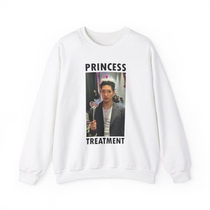 Może przedstawiać: Biała bluza z nadrukiem zdjęcia osoby w koronie i z różdżką. Słowa "PRINCESS TREATMENT" są wydrukowane w kolorze czarnym nad i pod zdjęciem. Bluza ma okrągły dekolt.