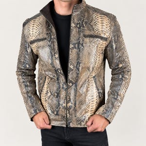 May include: A jacket featuring a tan, brown, and black snakeskin pattern with a stand-up collar, a full-length front zipper, and two horizontal zippered chest pockets. The garment is worn over a solid black shirt and paired with dark pants.