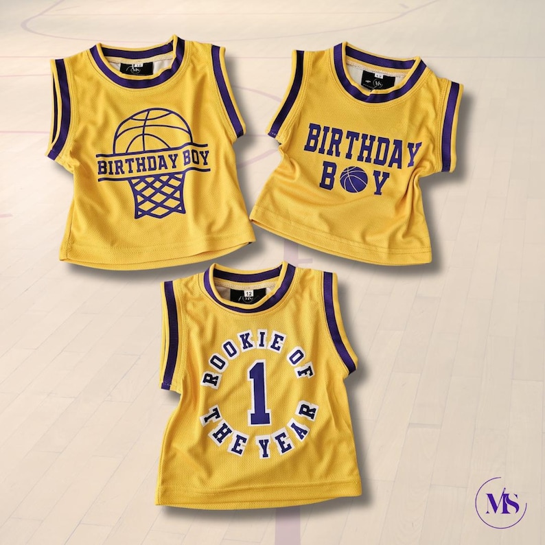 Puede incluir: Tres camisetas de baloncesto amarillas sin mangas con ribetes morados. La primera camiseta tiene un gr&aacute;fico de baloncesto y red con el texto "Birthday Boy". La segunda camiseta tiene el texto "Birthday Boy" con un gr&aacute;fico de baloncesto. La tercera camiseta tiene el texto "Rookie of the Year" con el n&uacute;mero "1" en un c&iacute;rculo.