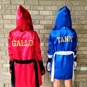 Puede incluir: Dos batas de boxeo de satén rojo y azul con capucha. La bata roja tiene el nombre "GALLO" bordado en la espalda con letras doradas. La bata azul tiene el nombre "TANK" bordado en la espalda con letras doradas.