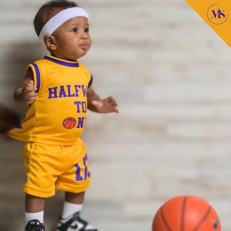 Puede incluir: Un ni&ntilde;o peque&ntilde;o vistiendo una camiseta de baloncesto amarilla y morada con el texto "Halfway to One" y un gr&aacute;fico de baloncesto. El ni&ntilde;o lleva una diadema blanca y sostiene un bal&oacute;n de baloncesto.