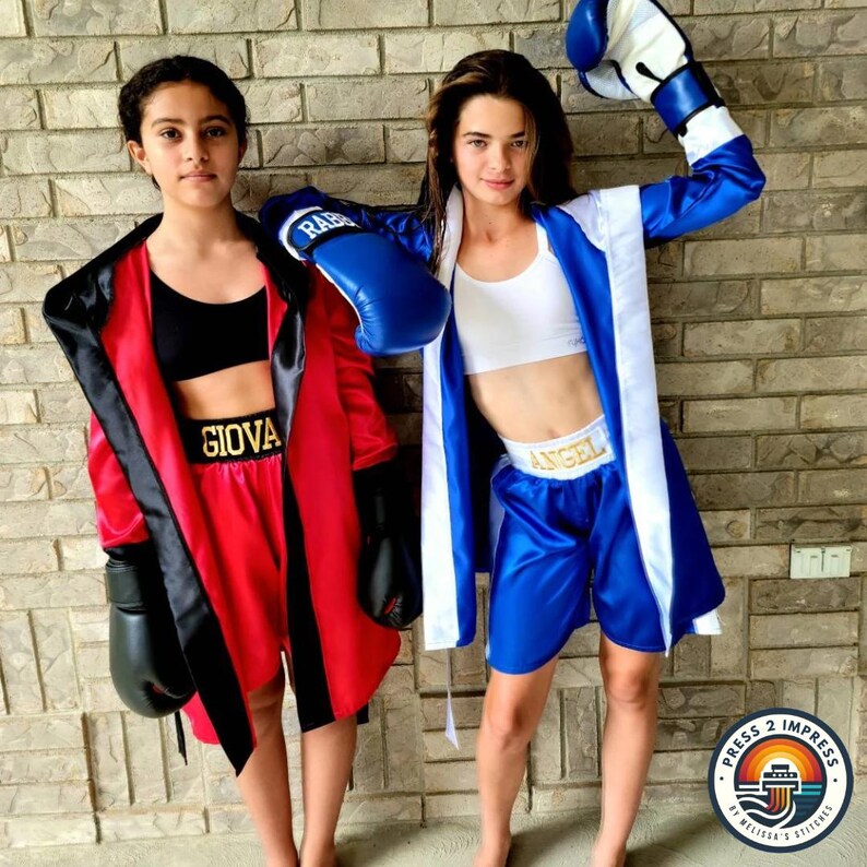 May include: Two girls wearing boxing robes and shorts. One girl is wearing a red robe with black trim and black boxing gloves. The other girl is wearing a blue robe with white trim and blue boxing gloves. Both girls are wearing gold belts with their names on them. The girl in the red robe has the name "Giova" on her belt. The girl in the blue robe has the name "Angel" on her belt.