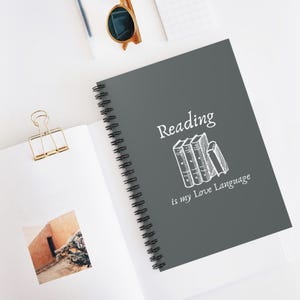 May include: A grey spiral notebook with the text "Reading is my Love Language" in white. The text is accompanied by a drawing of a stack of books.
