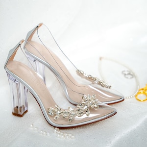 Può includere: Un paio di scarpe con tacco alto trasparenti con un ornamento di glitter argentato sulla punta. Le scarpe hanno un tacco a spillo trasparente e una punta a punta.