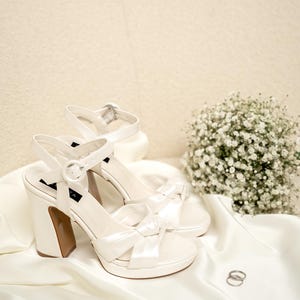 Puede incluir: Un par de zapatos de novia de sat&eacute;n blanco con un detalle de lazo y un tac&oacute;n blanco grueso. Los zapatos est&aacute;n sobre una tela de sat&eacute;n blanco con dos anillos plateados.