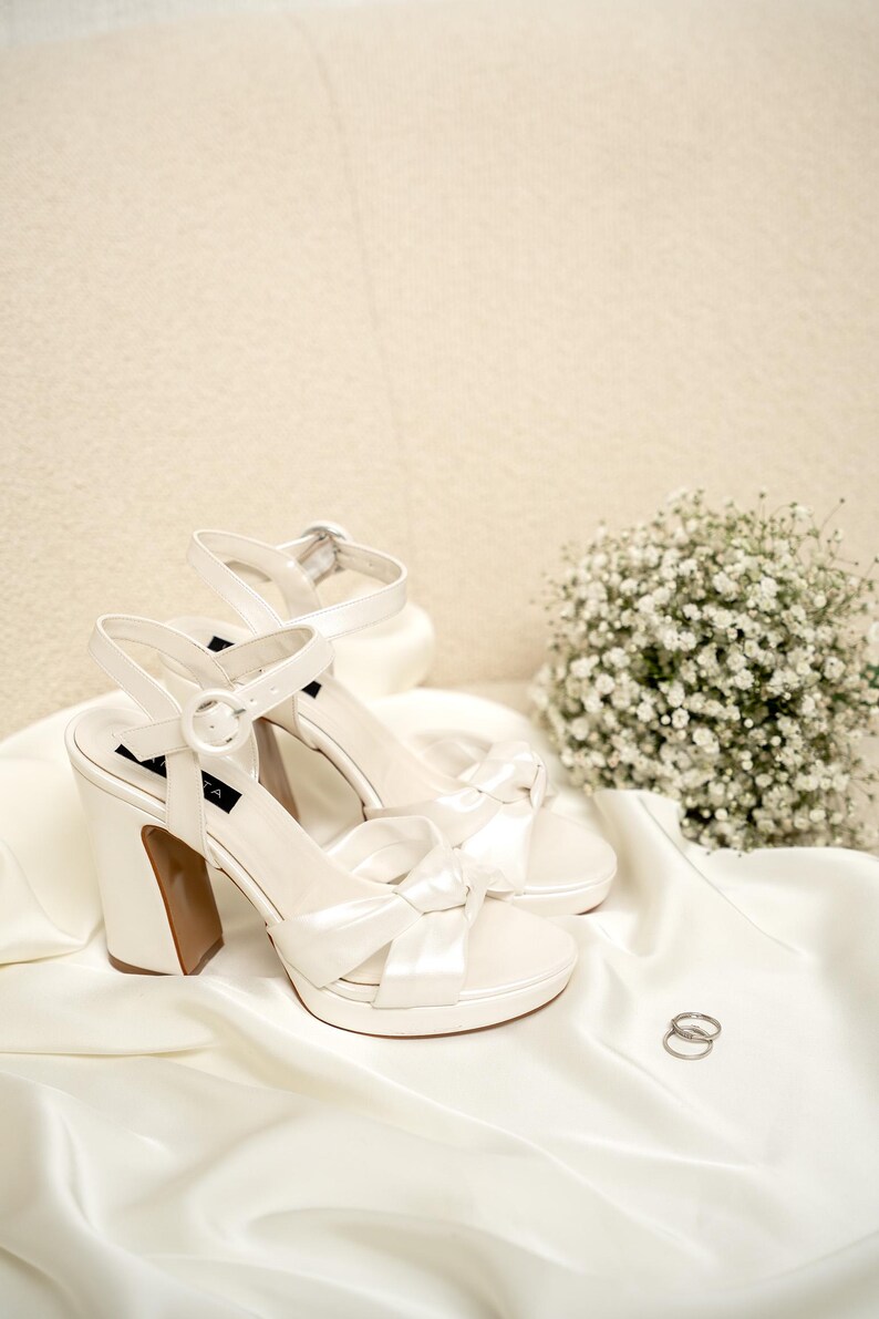 Puede incluir: Un par de zapatos de novia de sat&eacute;n blanco con un detalle de lazo y un tac&oacute;n blanco grueso. Los zapatos est&aacute;n sobre una tela de sat&eacute;n blanco con dos anillos plateados.