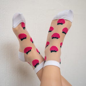 May include: A pair of white sheer socks with a pink strawberry pattern. The socks are worn on a person's legs, with the feet crossed.
