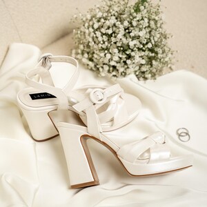 Puede incluir: Un par de tacones de novia de sat&eacute;n blanco con un tac&oacute;n grueso y una correa de tobillo. Los tacones est&aacute;n sobre una superficie de tela blanca con un ramo de aliento de beb&eacute; blanco.