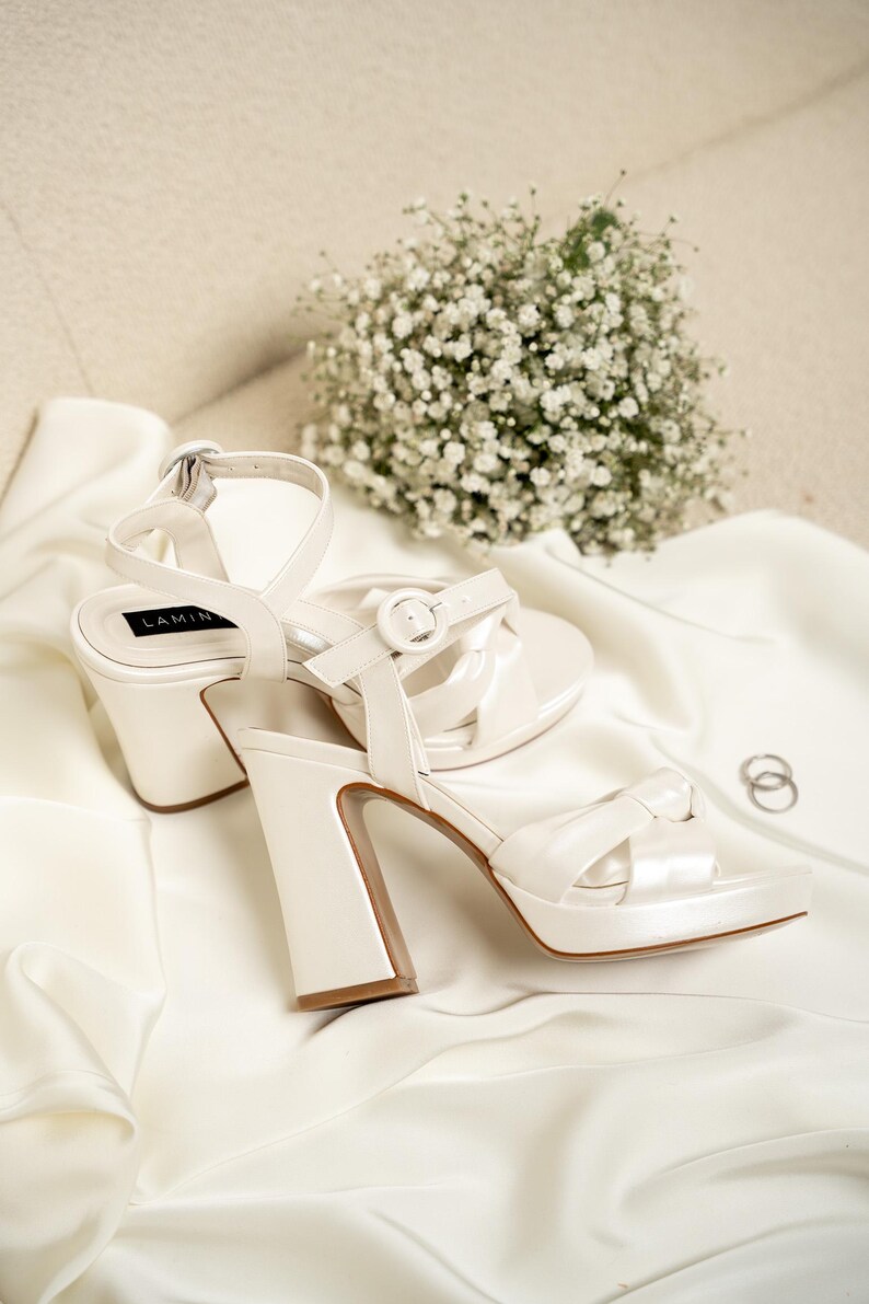 Puede incluir: Un par de tacones de novia de sat&eacute;n blanco con un tac&oacute;n grueso y una correa de tobillo. Los tacones est&aacute;n sobre una superficie de tela blanca con un ramo de aliento de beb&eacute; blanco.