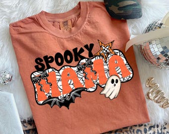 May include: An orange Comfort Colors t-shirt with the words "Spooky Mama" in black and orange, featuring a bat, spiderwebs, a star, and a ghost graphic. A camouflage baseball cap with the word "LOVE ME" is visible in the background.