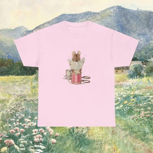 Pode incluir: Uma t-shirt rosa claro com um gráfico central de um rato castanho sentado num carretel de linha vermelho, a ler um jornal. O rato está rodeado por um pequeno dedal e uma tesoura de metal, tudo num estilo de ilustração suave.