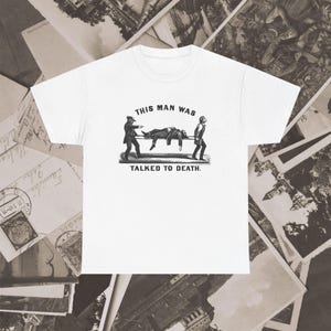 以下が含まれることがあります： 白い半袖Tシャツで、担架に乗せられた人を運ぶ2人の人物を描いたモノクロのヴィンテージ風イラストがプリントされています。グラフィックの上下には、THIS MAN WAS TALKED TO DEATH.というテキストが記されています。