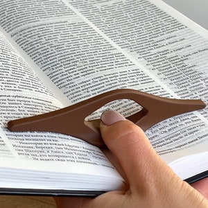 May include: A brown bookmark with a curved design is being used to hold open a book. The book is open to a page with text in a foreign language. The bookmark is being held by a hand with a light brown nail polish.