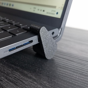 以下が含まれることがあります： 黒いラップトップコンピューターが、黒いラップトップスタンドの上に置かれています。ラップトップは開いており、画面にはウェブサイトが表示されています。スタンドは、テクスチャ加工された黒いプラスチックでできています。