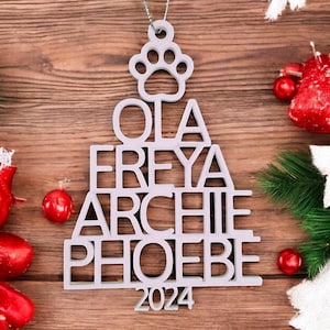 May include: A light gray Christmas tree ornament with a paw print at the top and the names OLA, FREYA, ARCHIE, and PHOEBE, along with the year 2024. The ornament hangs against a wooden background, decorated with red ornaments and greenery.