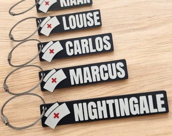 Puede incluir: Cinco llaveros negros con texto blanco y una cruz roja sobre una gorra de enfermera blanca. Los nombres en los llaveros son: Kian, Louise, Carlos, Marcus y Nightingale.