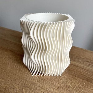 Può includere: Un vaso bianco, di forma quadrata, con un design ondulato e strutturato. Il vaso ha un'ampia apertura ed è realizzato in un materiale opaco e solido. L'esterno presenta linee verticali ondulate, creando un'estetica moderna. Il vaso è su una superficie di legno.