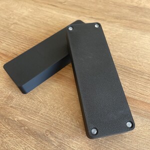 May include: Two black rectangular boxes with rounded edges and silver screws on the bottom.